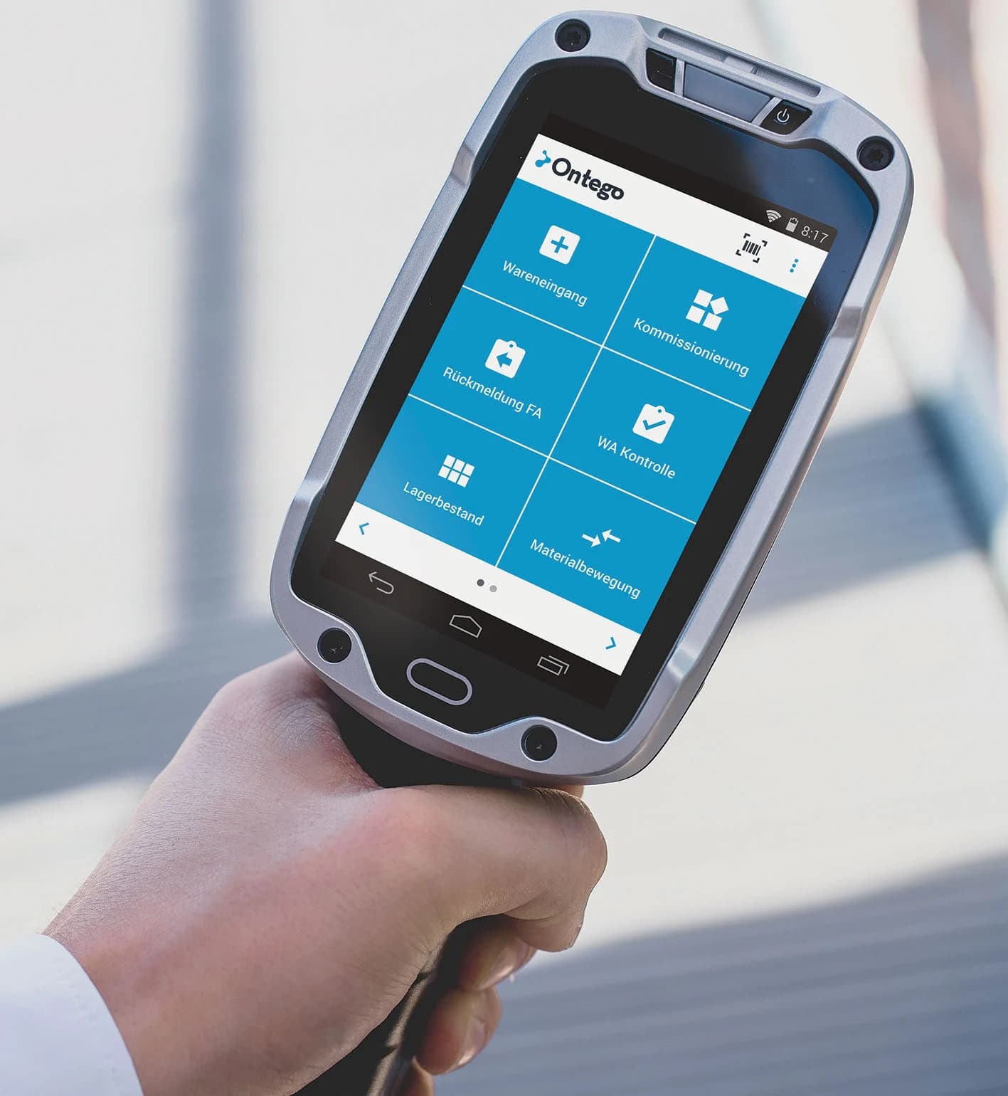 A close-up of a hand holding a rugged mobile device displaying the Ontego logistics app menu with various warehouse functions in German.