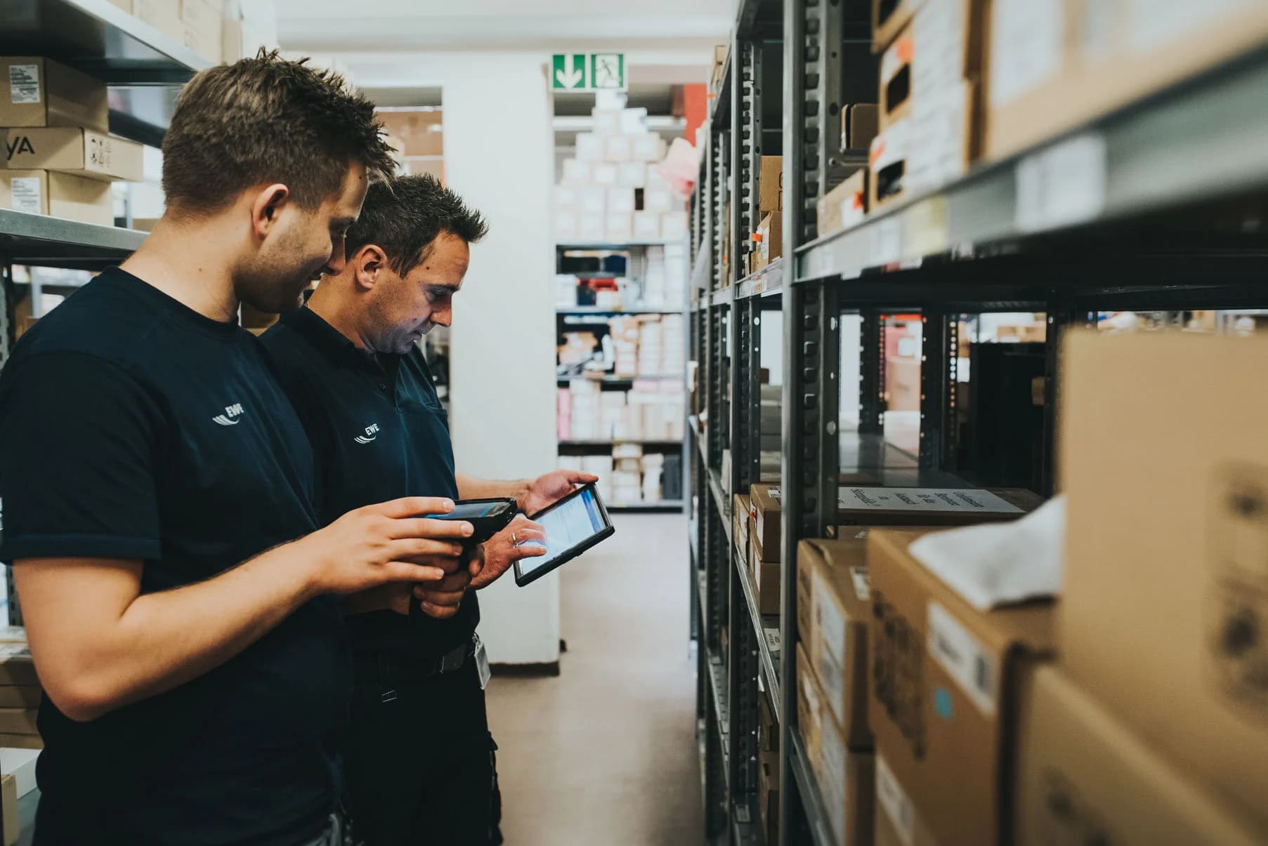 Two warehouse workers in dark uniforms using handheld devices and a tablet to manage inventory in a storage aisle filled with labeled cardboard boxes.