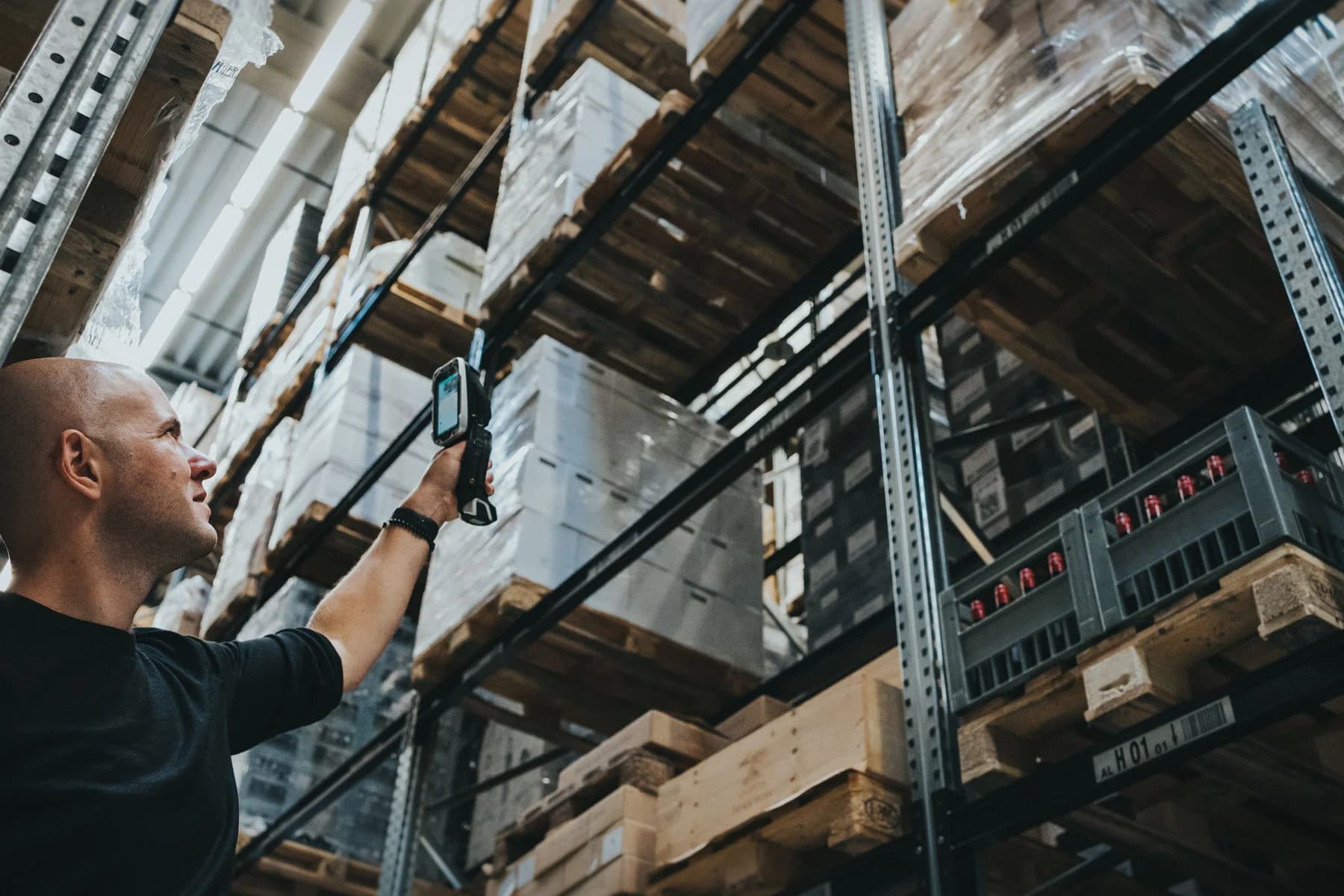 A man stands in a warehouse and lifts a mobile handheld device with an Ontego screen to scan a pallet stored on a high shelf. Stacked goods on pallets surround him.