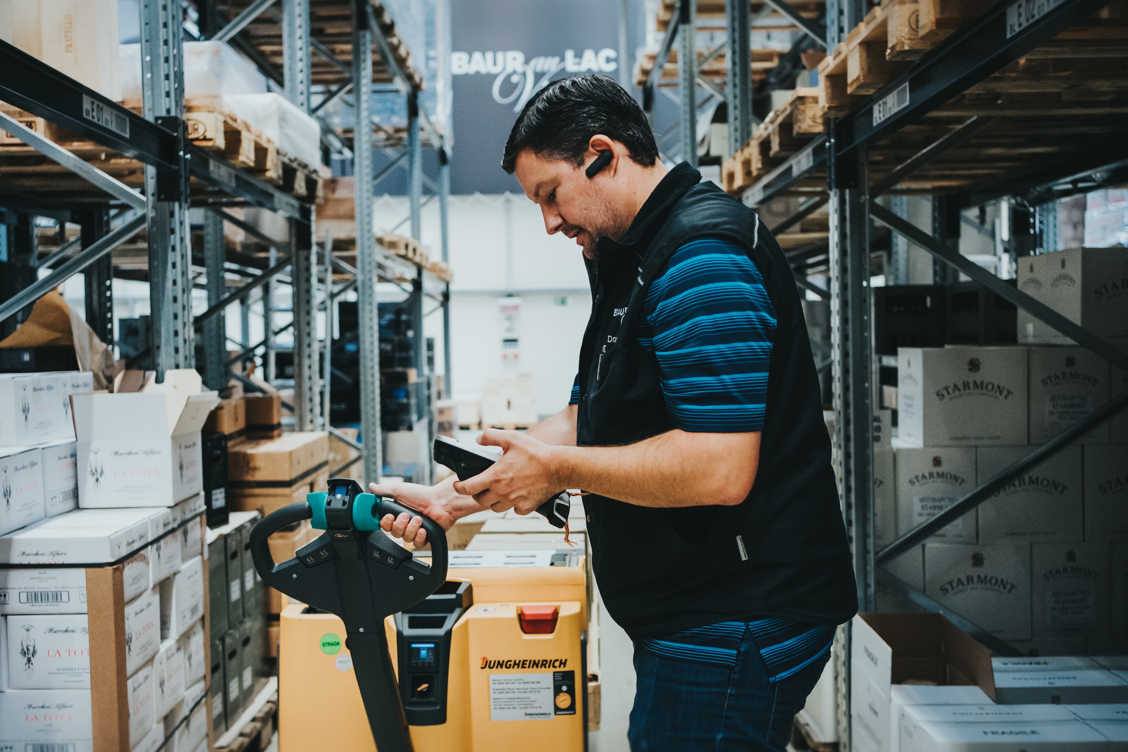 Warehouse worker using a handheld scanner during SAP supported picking, standing between storage racks with a pallet truck in a logistics environment.
