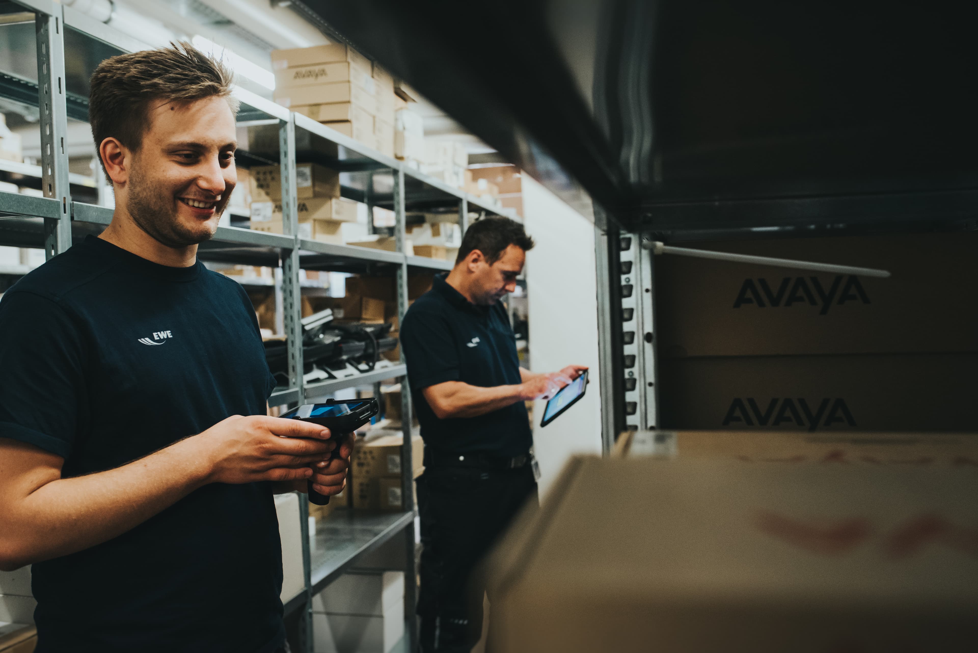 Two warehouse employees using mobile scanning devices among shelves stocked with boxes, illustrating digital inventory management and mobile data collection.
