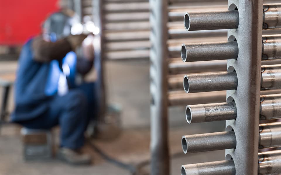 Close-up of parallel metal tubes in a heat exchanger with a blurred worker in blue safety clothing visible in the background at an industrial facility