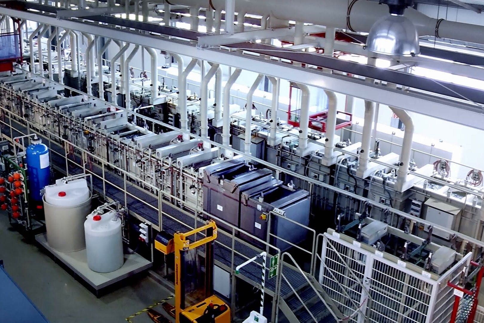 Modern industrial manufacturing facility with multiple production lines, featuring white structural columns, automated machinery, chemical storage tanks, and a yellow automated guided vehicle on the factory floor