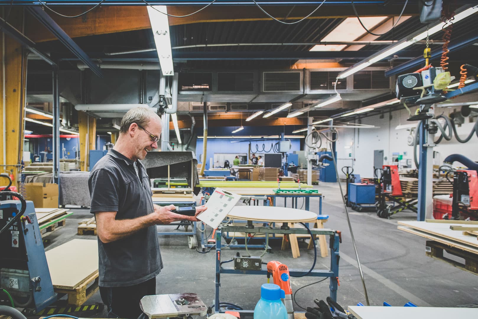 Worker in a production hall using a mobile scanner to record operation data, holding a job sheet beside machinery and workstations for SAP related time capture.