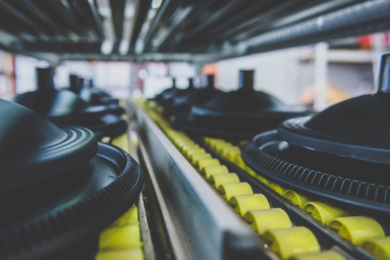 Rows of black rubber products with circular treaded patterns arranged on yellow support rails in a manufacturing facility, with blurred factory floor in the background