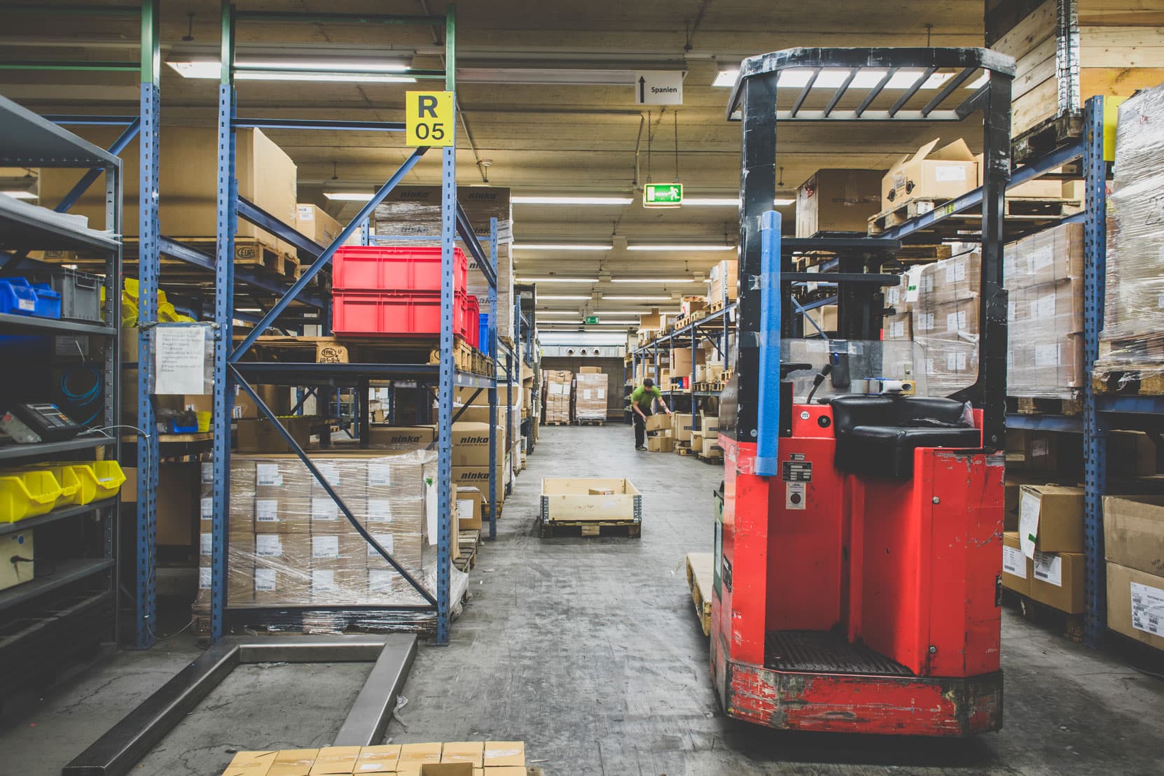 Warehouse aisle with shelves, packed pallets, forklift and worker organizing inventory